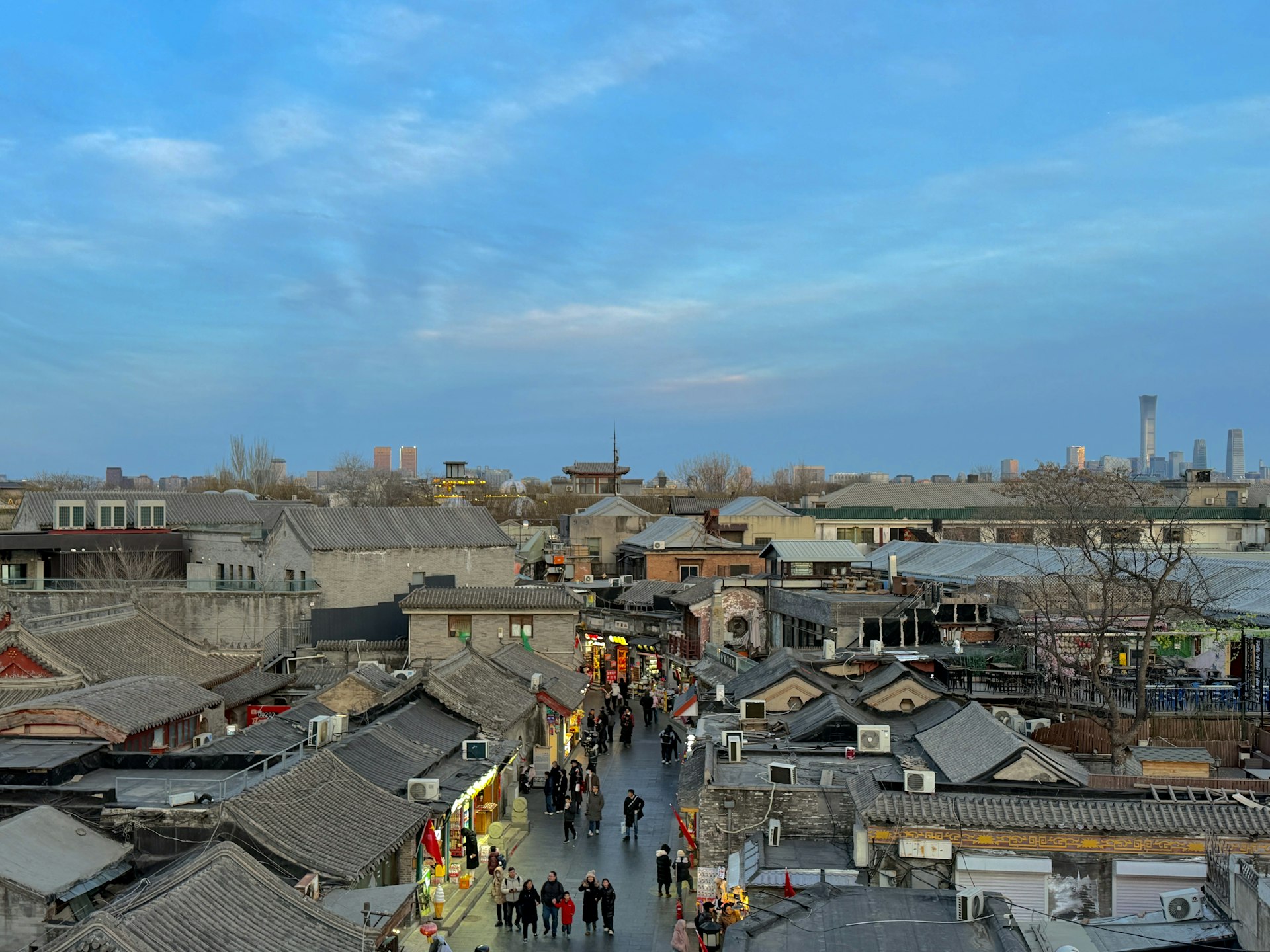A narrow Beijing hutong alley strung with red lanterns at dusk, a vendor grilling skewers in the foreground