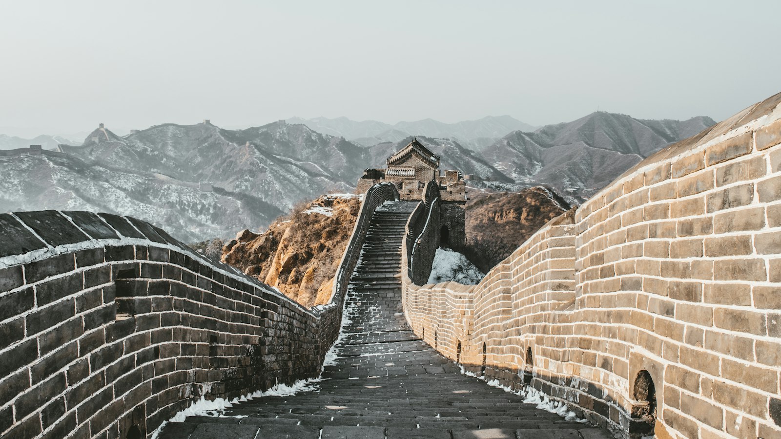 A snow-dusted stone walkway on the Great Wall climbing toward a watchtower, with snow-streaked mountains and more wall stretching into the haze beyond