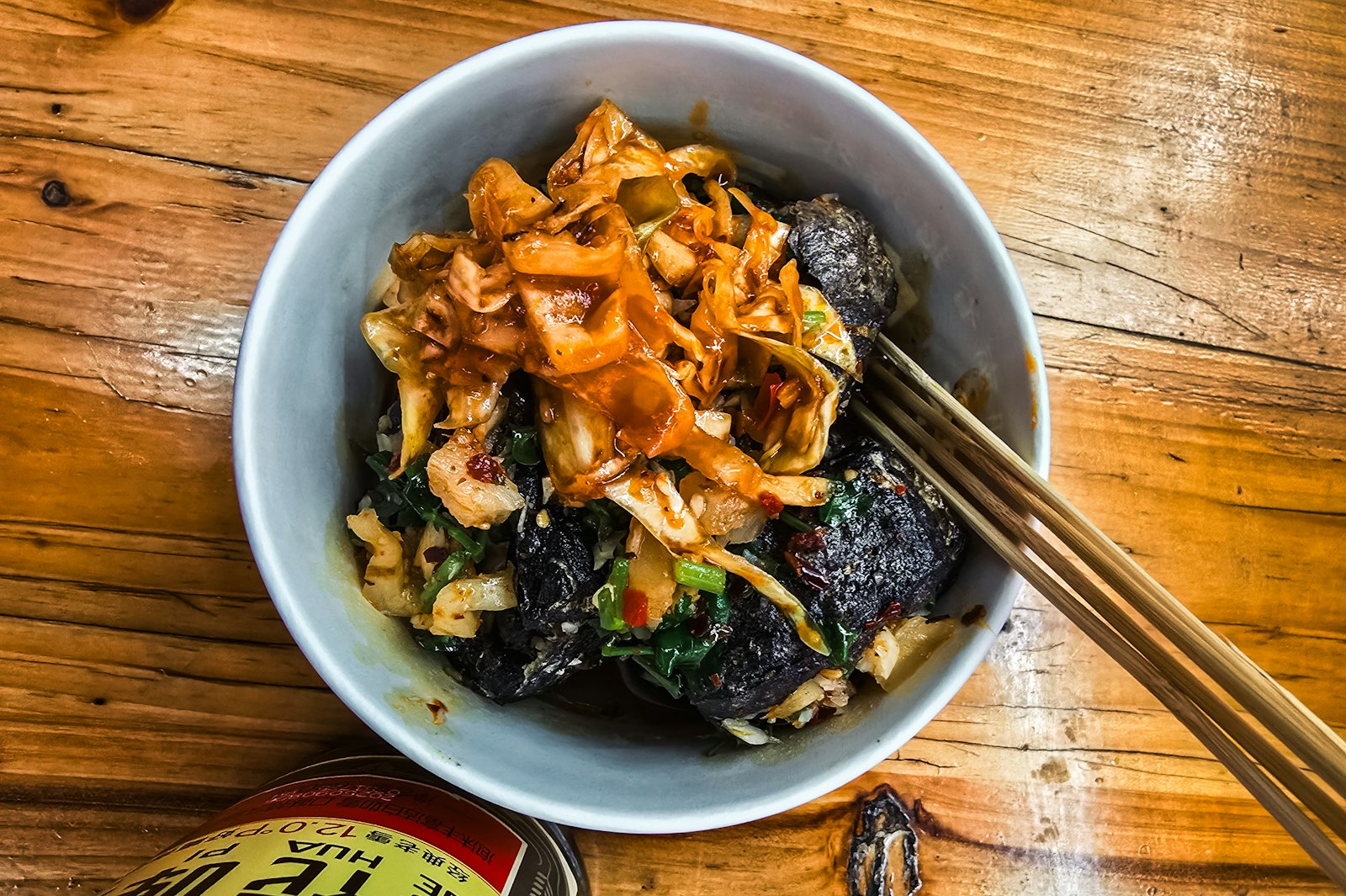 A bowl of black fried stinky tofu topped with shredded pickled cabbage, chili and scallions, with chopsticks resting on the rim