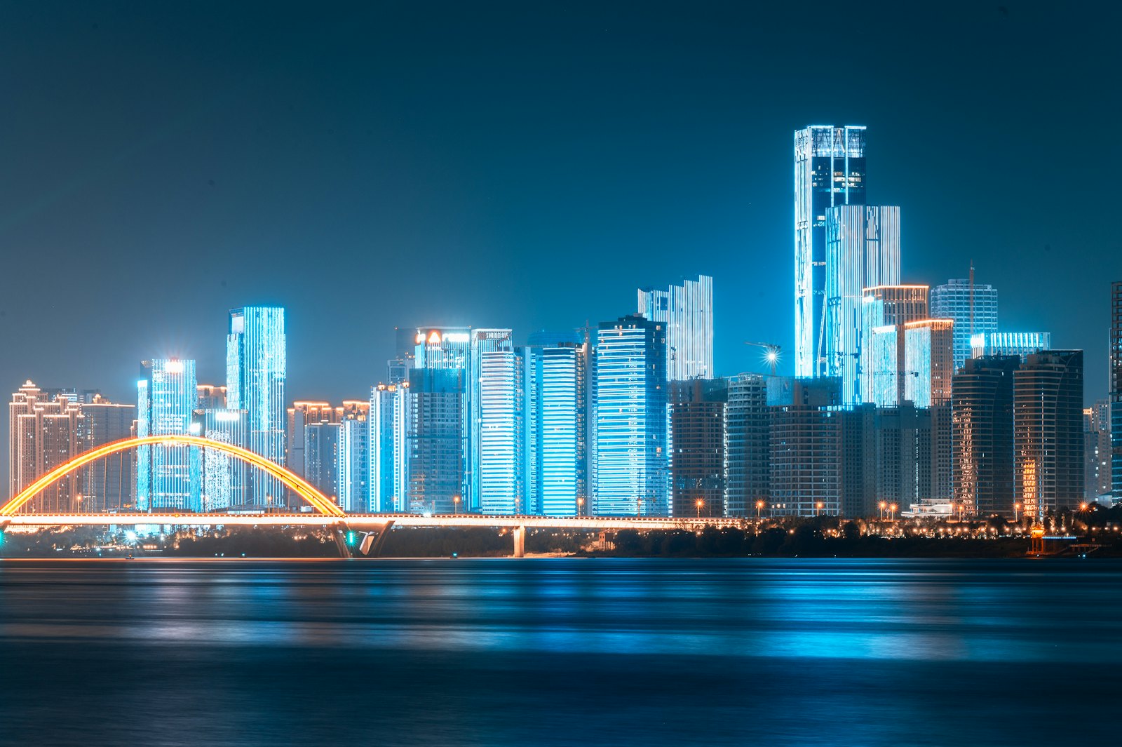 The Changsha skyline across the Xiang River at night, with towers lit in blue and an arched bridge outlined in yellow lights