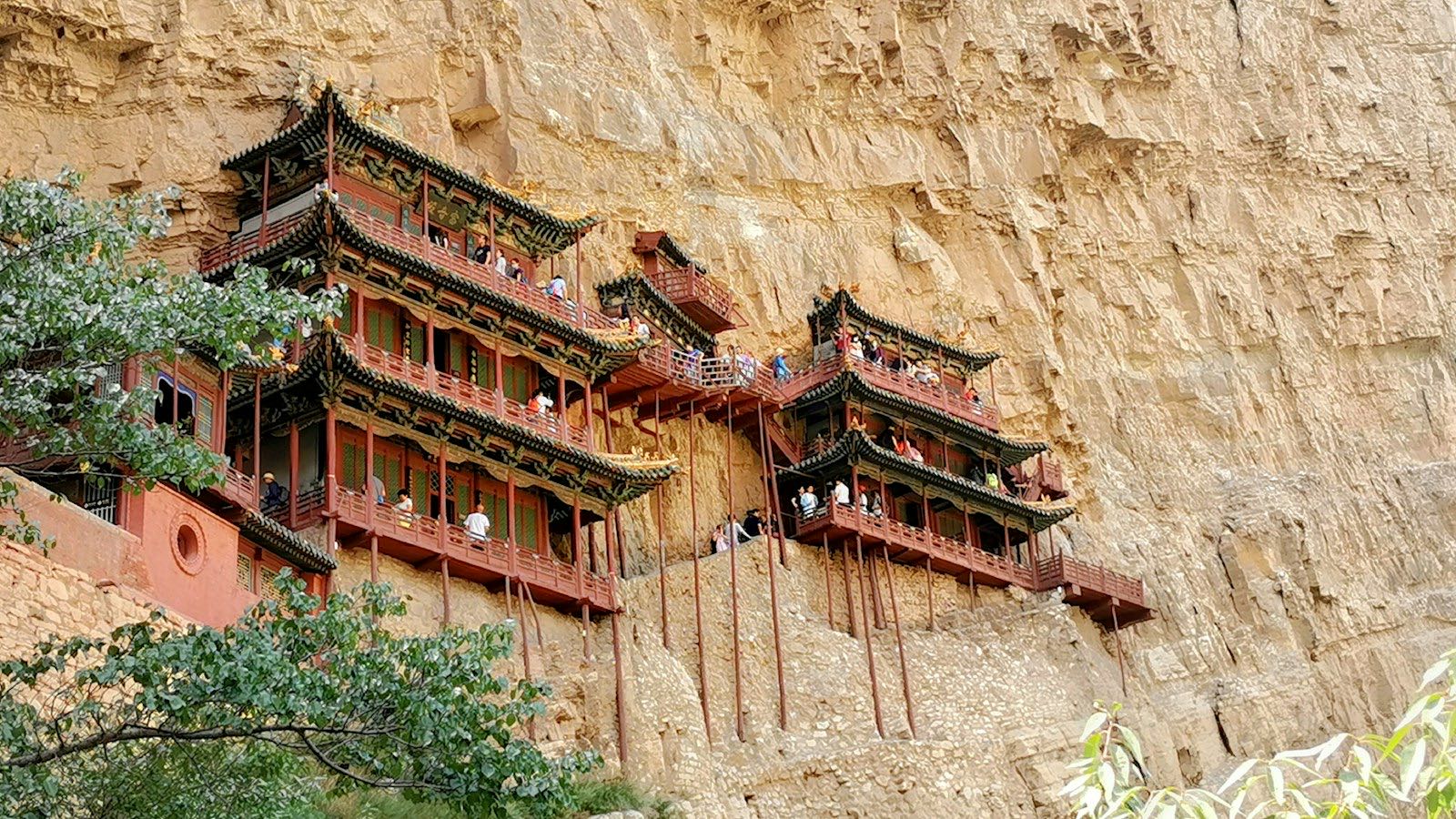 A closer look at the Hanging Temple: tiers of dark-tiled pavilions with red balustrades held up by slender wooden poles against the sandstone cliff, visitors visible on the upper walkways