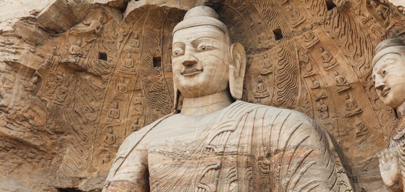 A colossal Buddha carved from the sandstone cliff at the Yungang Grottoes near Datong, with rows of tiny seated Buddhas carved into the wall behind him and part of a second figure at the right edge