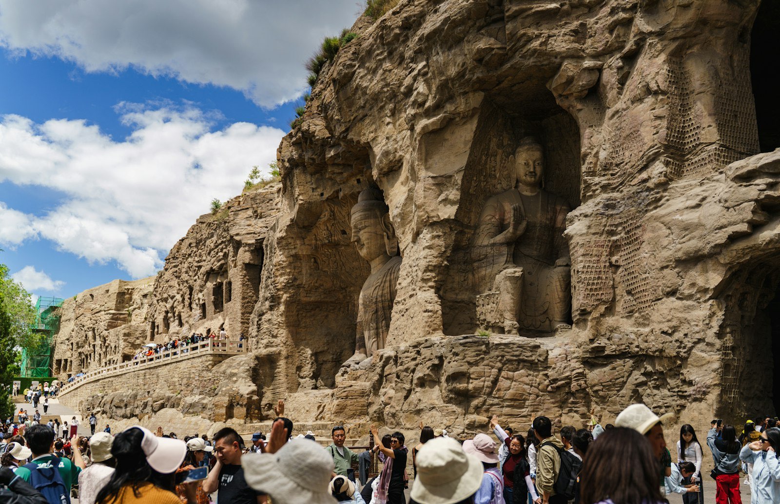 The Yungang Grottoes on a clear day with a blue sky and white clouds — a seated Buddha in an open niche in the sandstone cliff, a raised walkway along the rock face and a crowd of visitors in the foreground