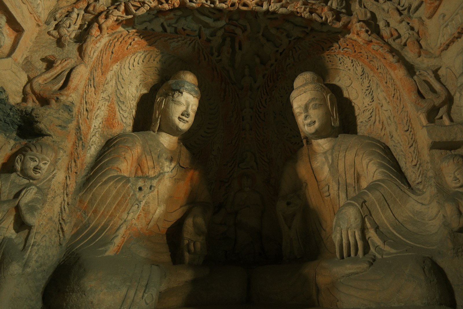 Two seated Buddhas face each other inside a carved niche at the Yungang Grottoes, the sandstone flecked with orange and a carved arch of tiny figures overhead
