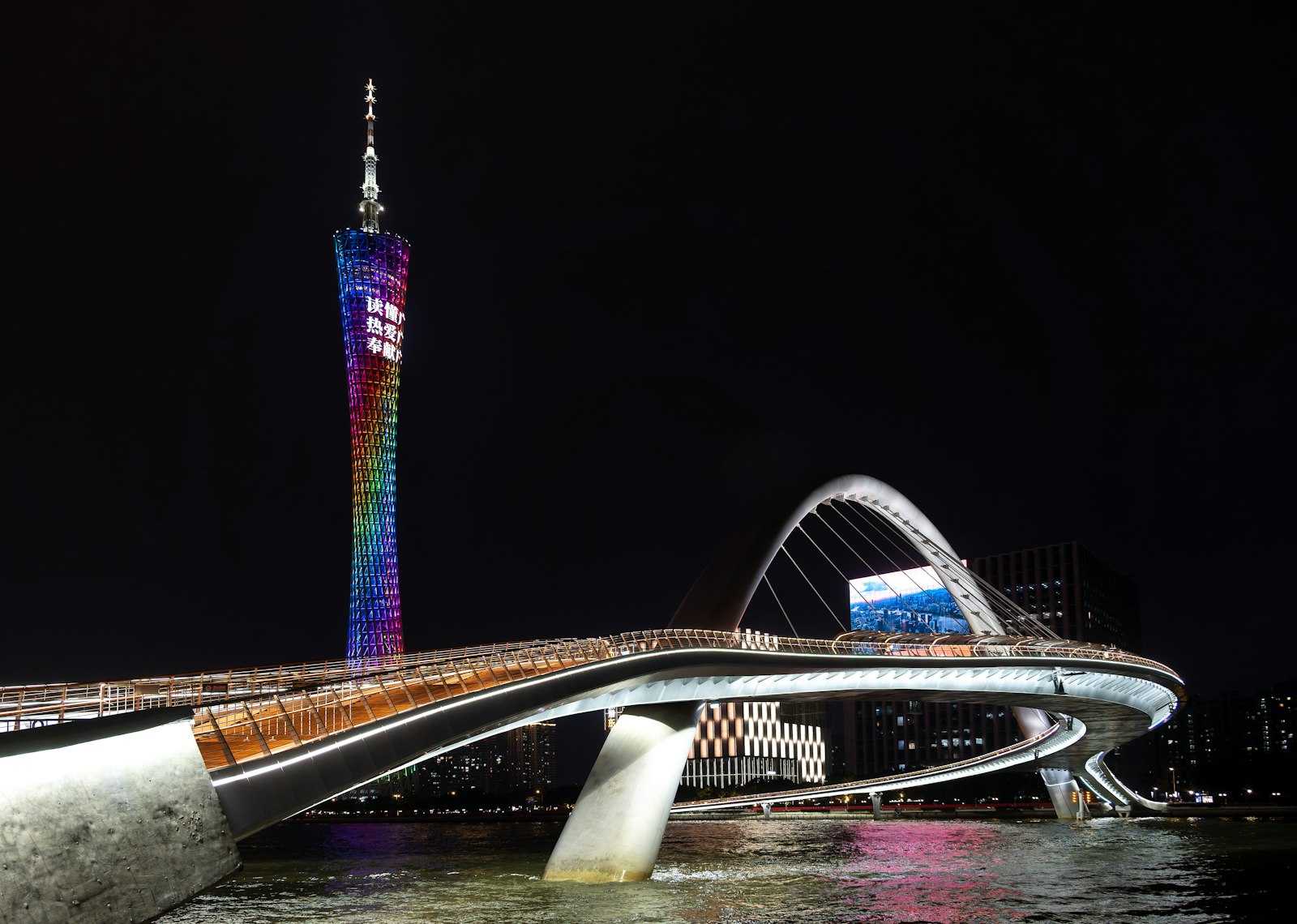 Canton Tower lit in rainbow colours after dark beyond a curving, white-lit pedestrian footbridge arching over the water