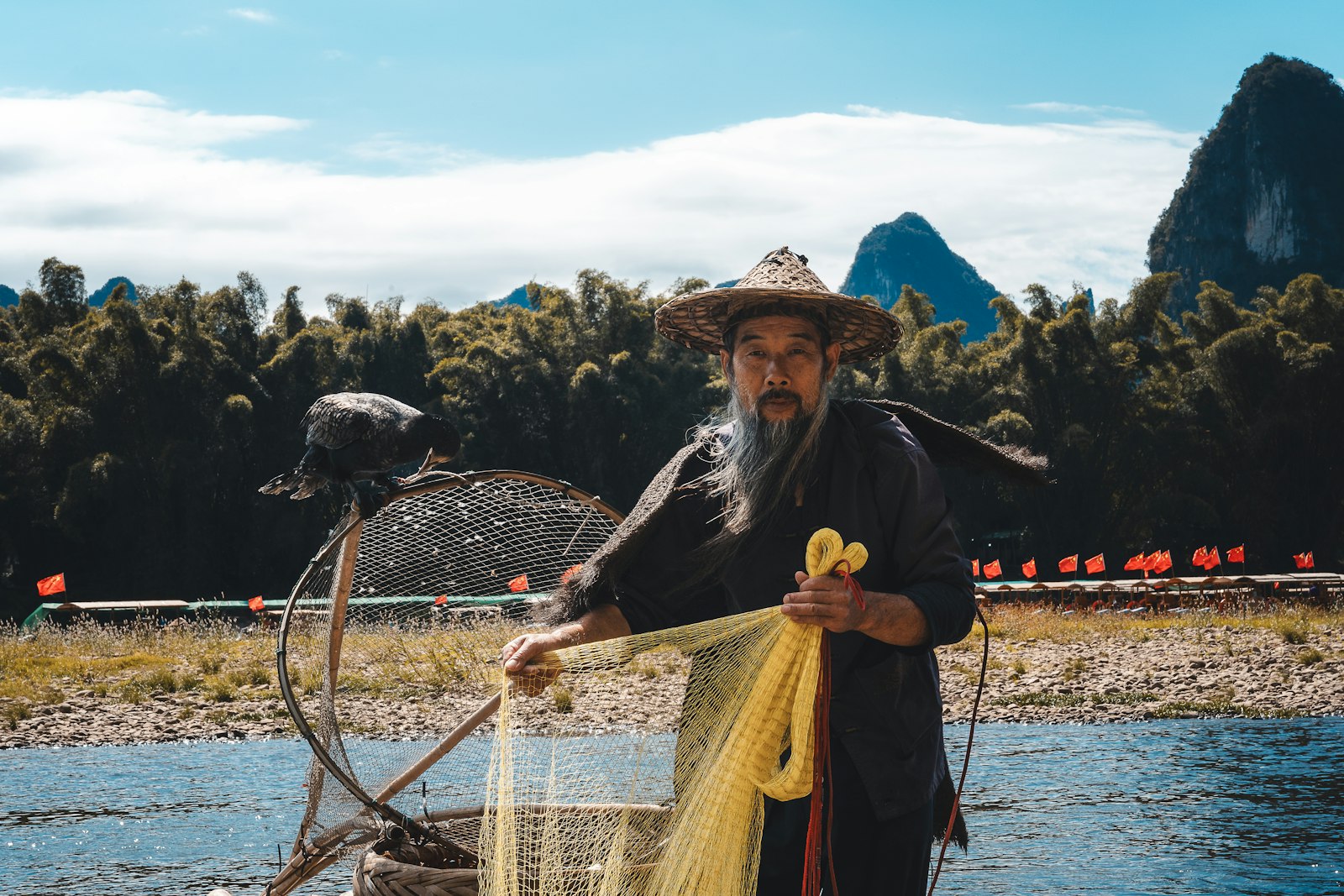 A Li River fisherman in a straw hat holding a yellow net, a cormorant perched on the rim of his hoop net, with bamboo groves and limestone peaks behind him