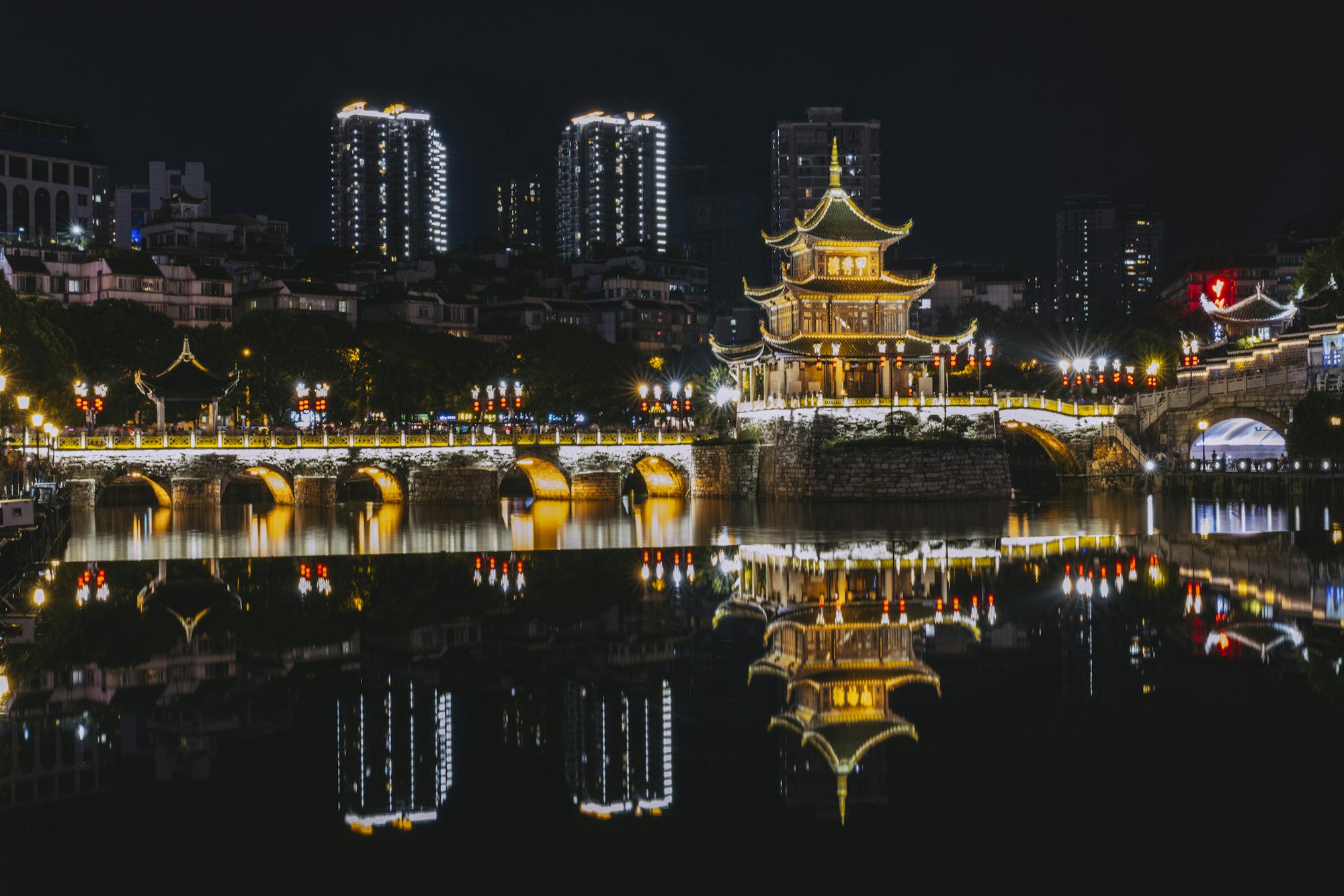 Jiaxiu Pavilion in Guiyang lit up in gold at night, its tiered roofs and the arched bridge beside it reflected in the Nanming River, with tall buildings behind