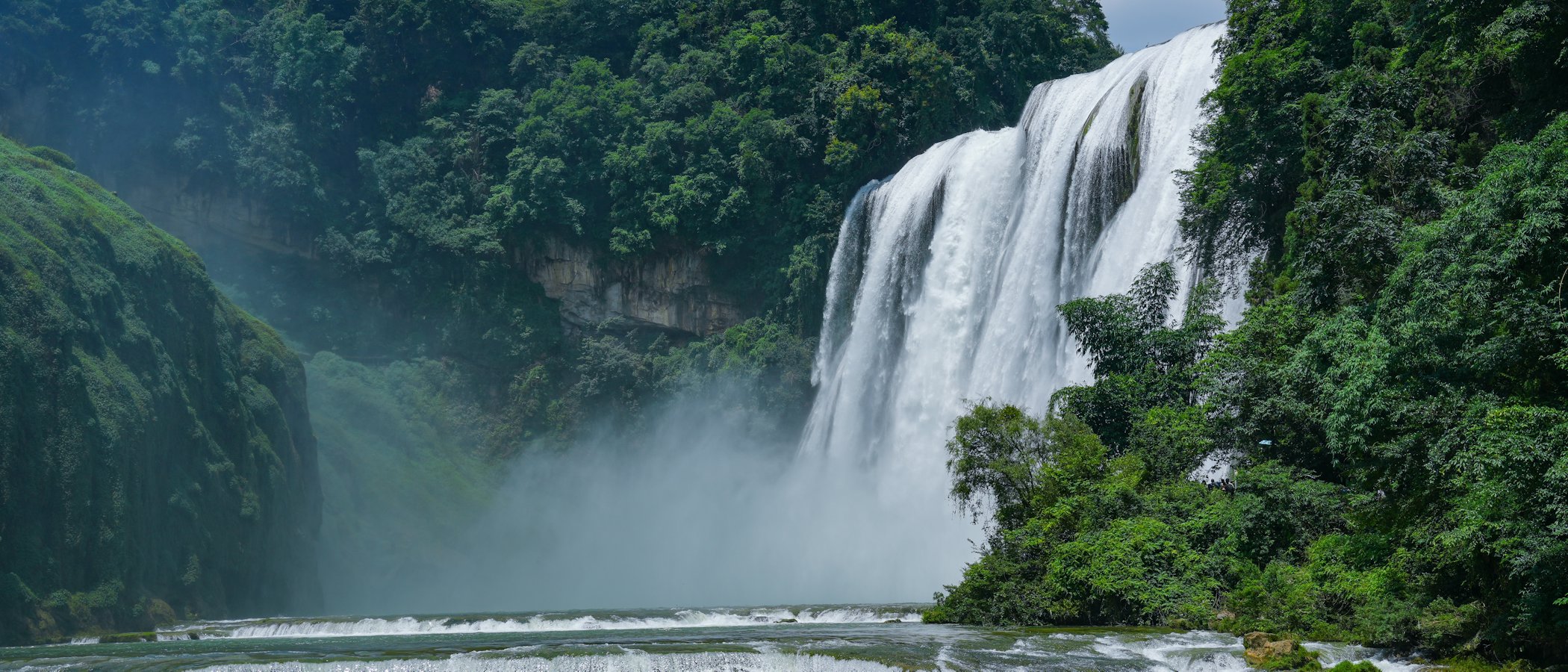 The Grand Waterfall at Huangguoshu, a tall white curtain of water pouring over a cliff into a mist-filled gorge, with a low stepped cascade in front and green forest on the slopes