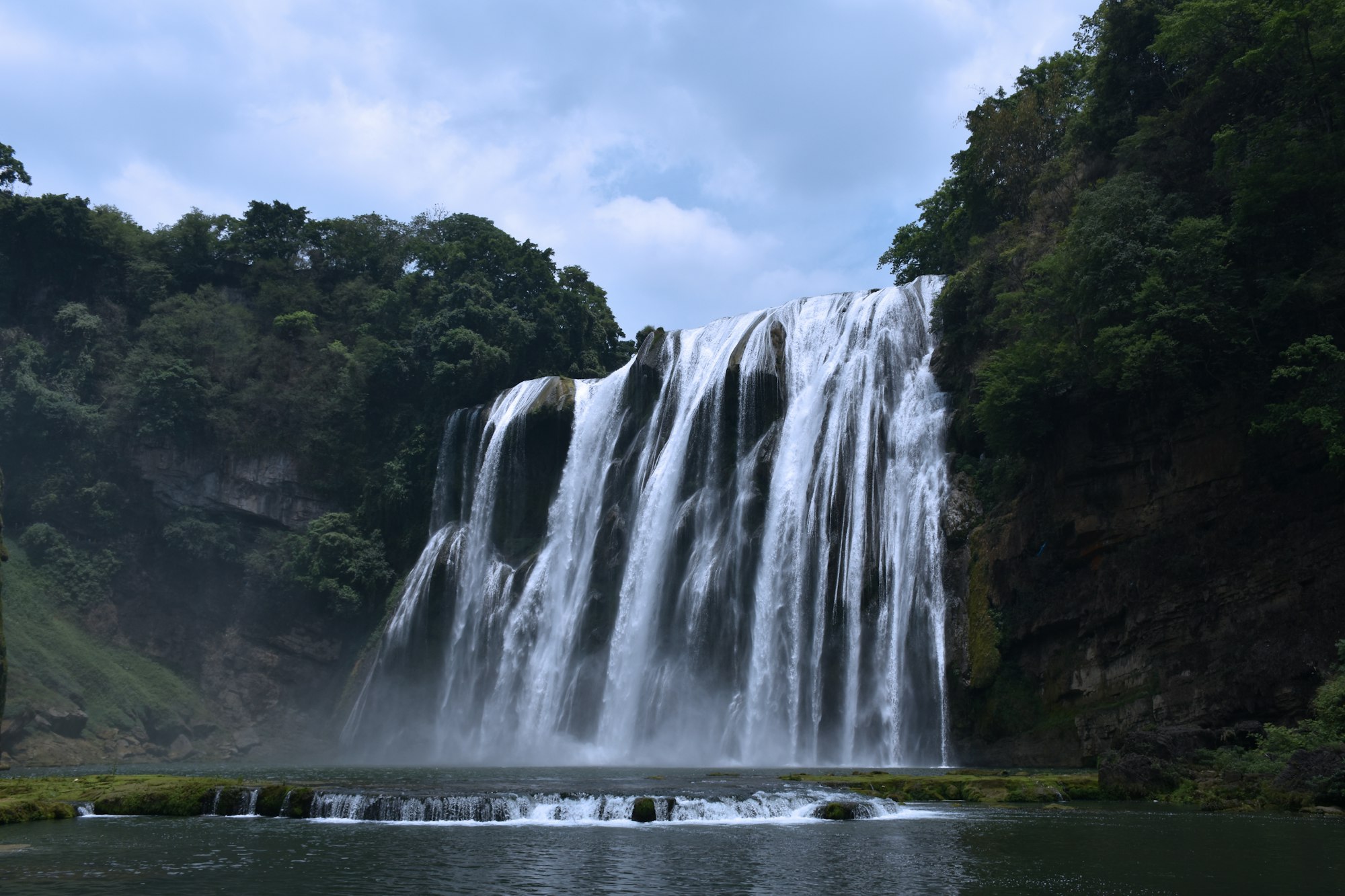 The Grand Waterfall at Huangguoshu, a wide white curtain of water pouring over a dark cliff into a green pool, with forest on both sides and a low cascade in front