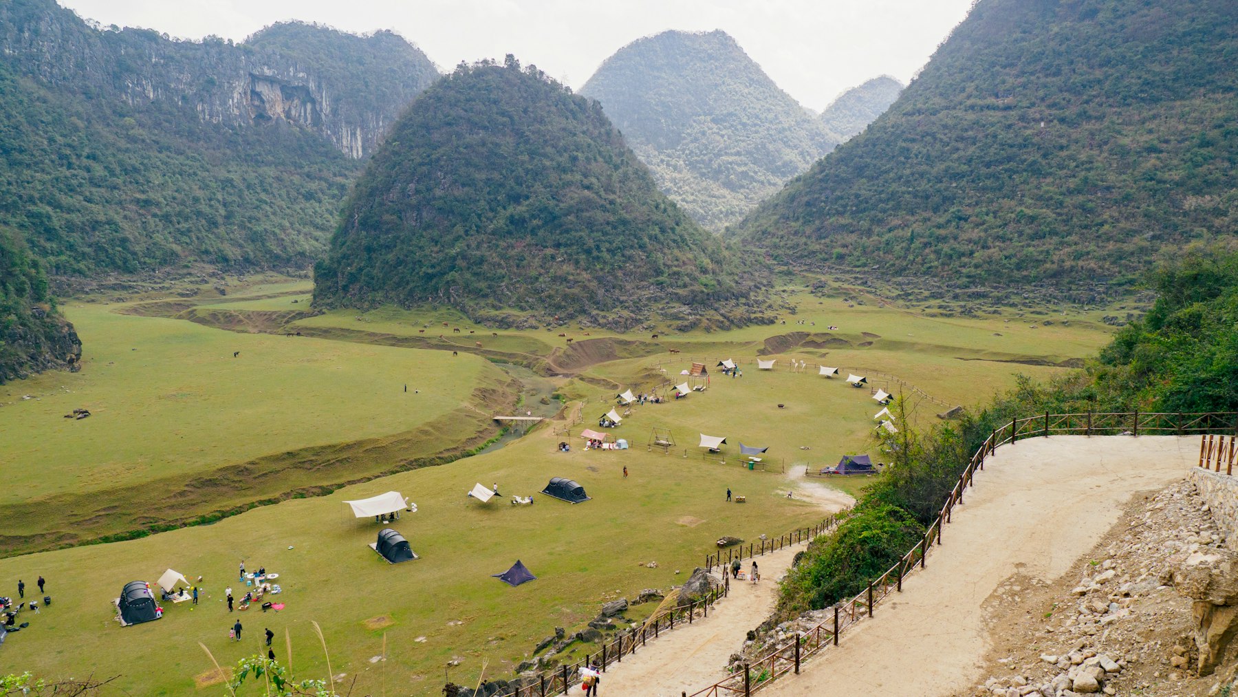 Green karst hills rising above a grassy valley dotted with small camping tents, with a dirt path and wooden fence in the foreground