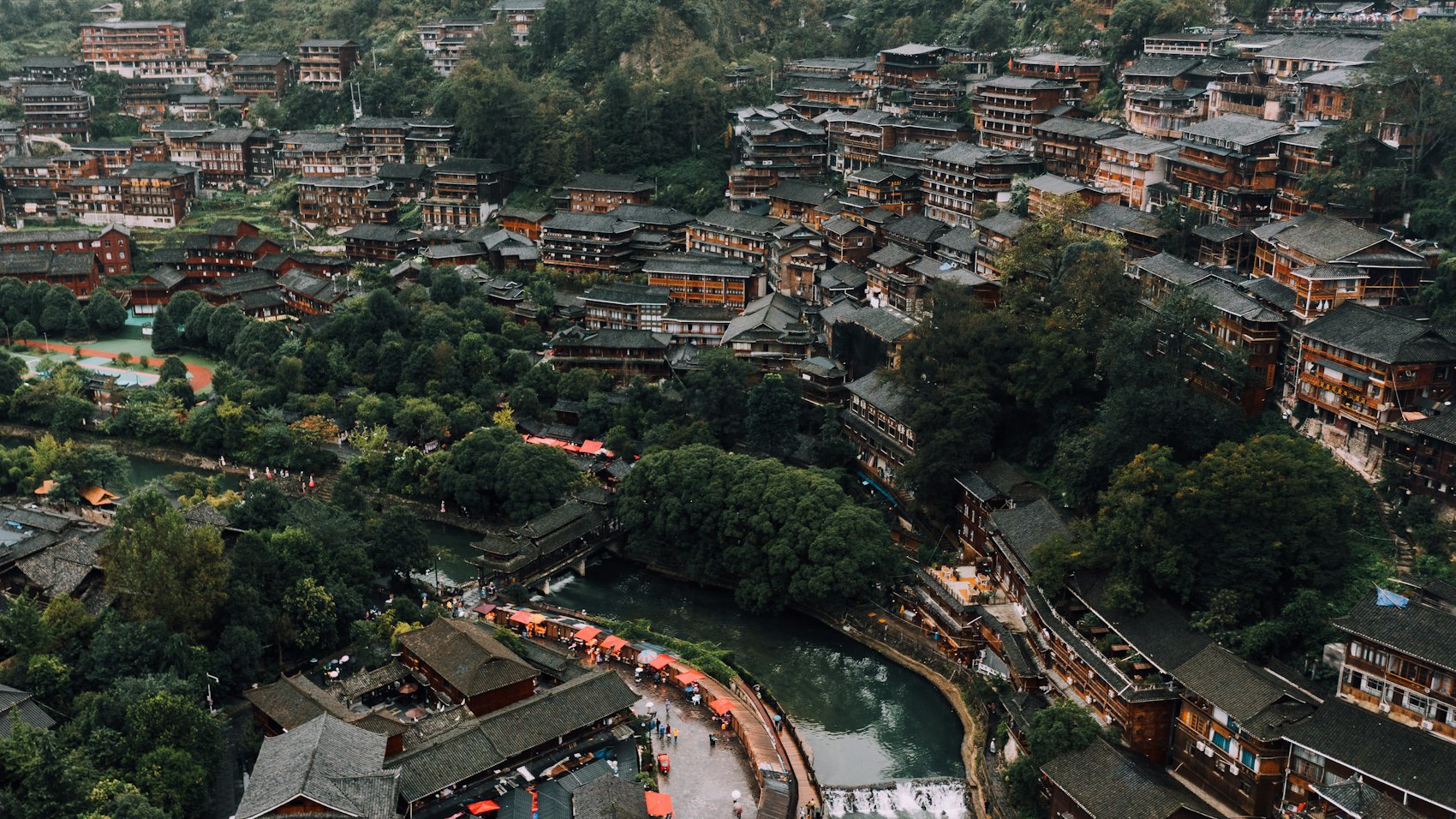 Aerial view of a hillside village of dark timber houses stacked above a river, with orange awnings along the bank and a small weir in the water
