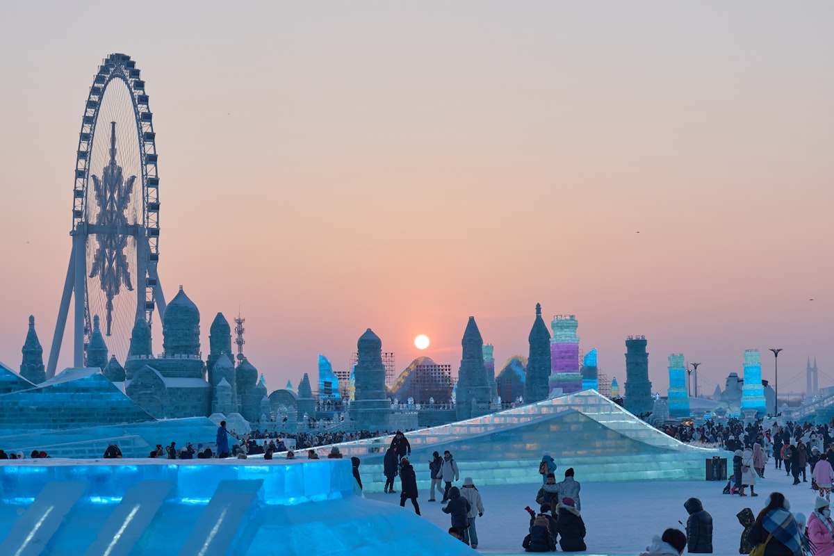 Harbin Ice and Snow World at night — illuminated ice castles and towers glowing blue, pink and green against the dark winter sky