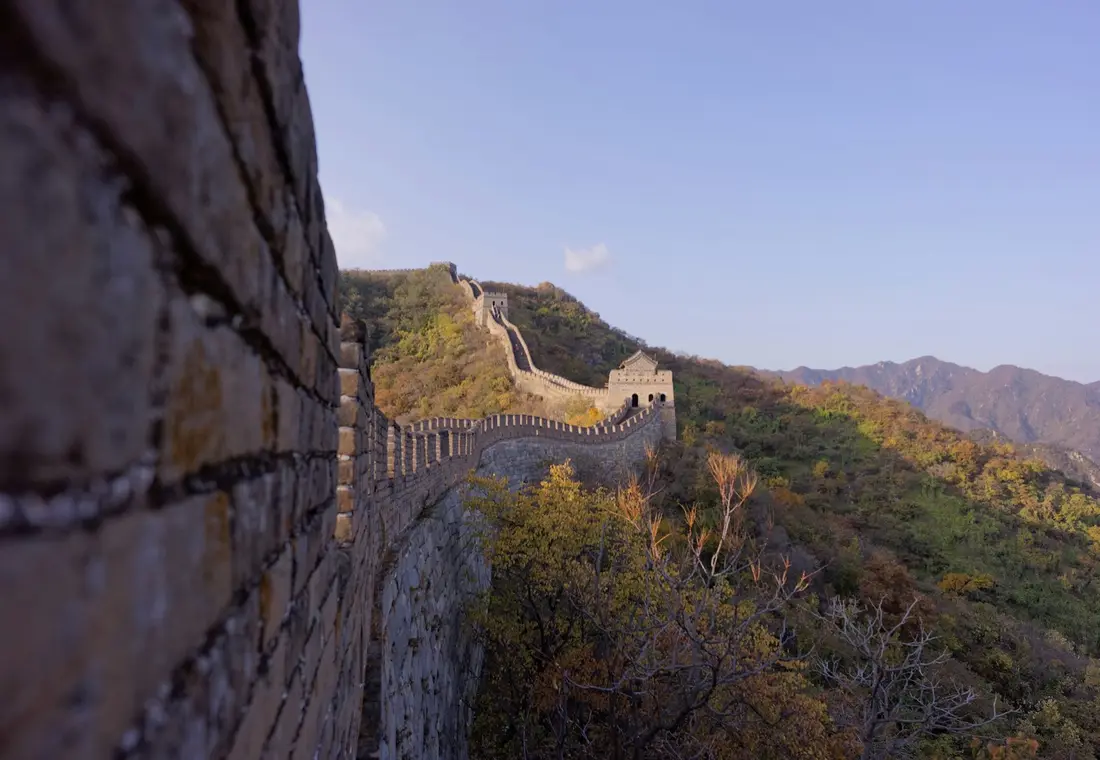 The Great Wall of China north of Beijing — a restored stretch of wall with crenellated parapets climbing a ridge past a watchtower, autumn-coloured hills and blue sky beyond
