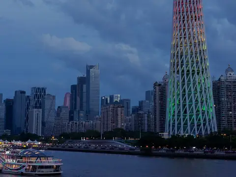 Canton Tower's twisting hourglass lattice lit in bands of red, orange, yellow and green at dusk above the Pearl River, with the city skyline behind and a lit-up cruise boat on the water in the foreground
