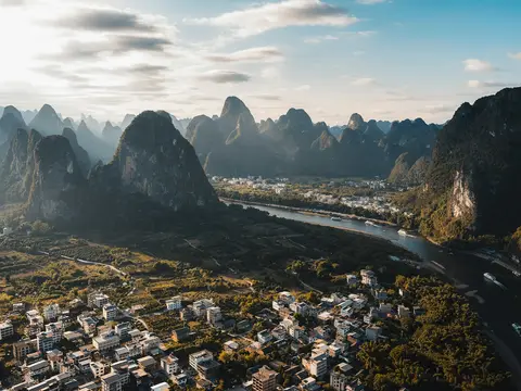 Aerial view of the Li River winding between towering limestone karst peaks in low golden sunlight, tourist boats on the water and a village of white houses on the bank