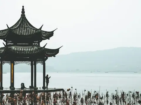 A two-tier wooden pavilion standing over West Lake in soft mist, dried lotus stalks in the water and hills fading into haze behind