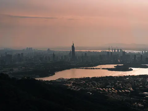 Nanjing skyline at sunset seen from Purple Mountain — Xuanwu Lake in the foreground, Zifeng Tower and the city's high-rises on a hazy horizon