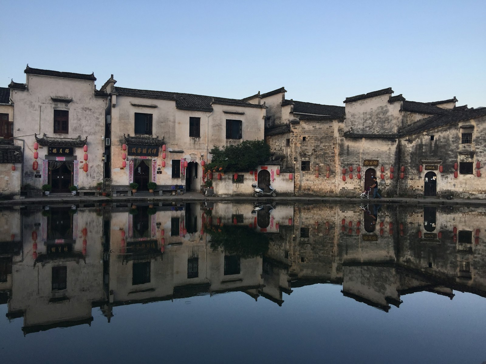 Hongcun in the early morning — weathered whitewashed Huizhou houses hung with red lanterns, mirrored in a perfectly still pond