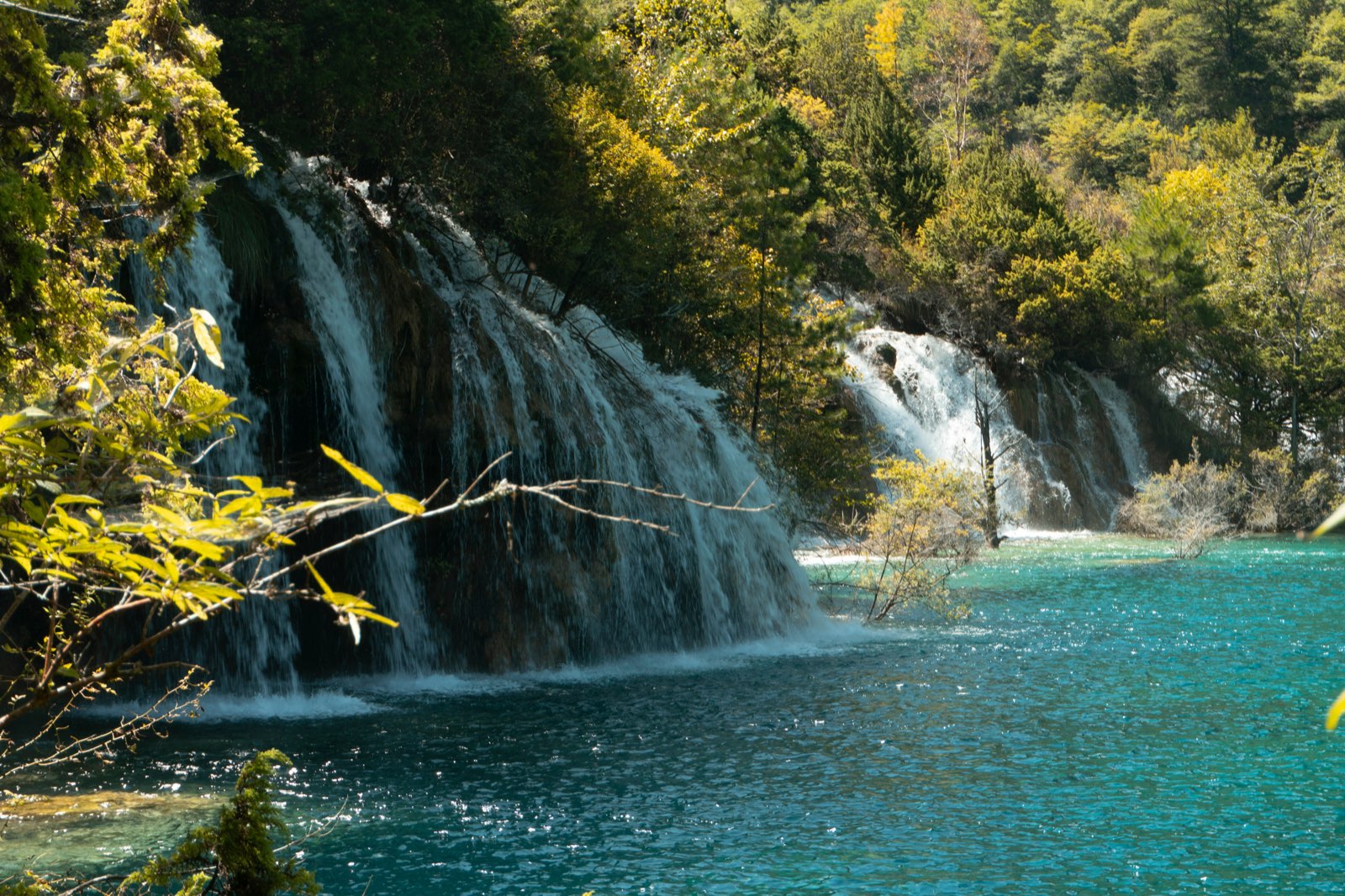 Waterfalls cascading down a rock face into a turquoise lake in Jiuzhaigou Valley, with yellow-green foliage around