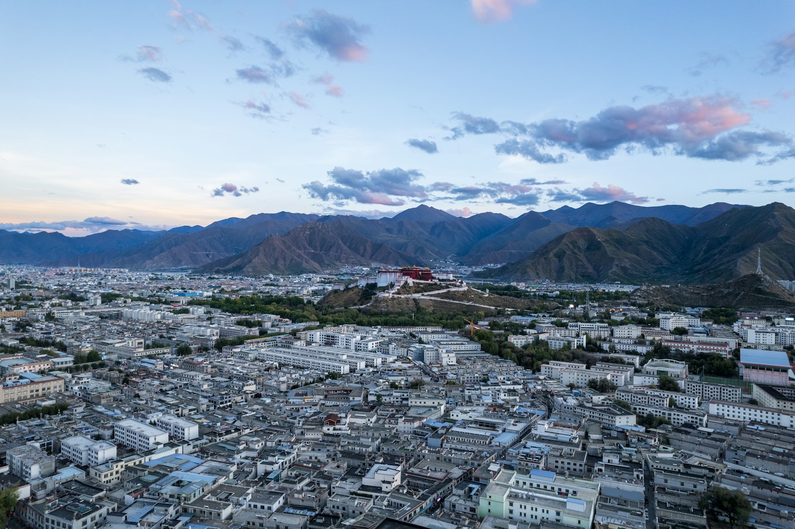 Aerial view of Lhasa at dusk — the city spread across its high valley, ringed by bare mountains, with the Potala Palace on its hill in the distance