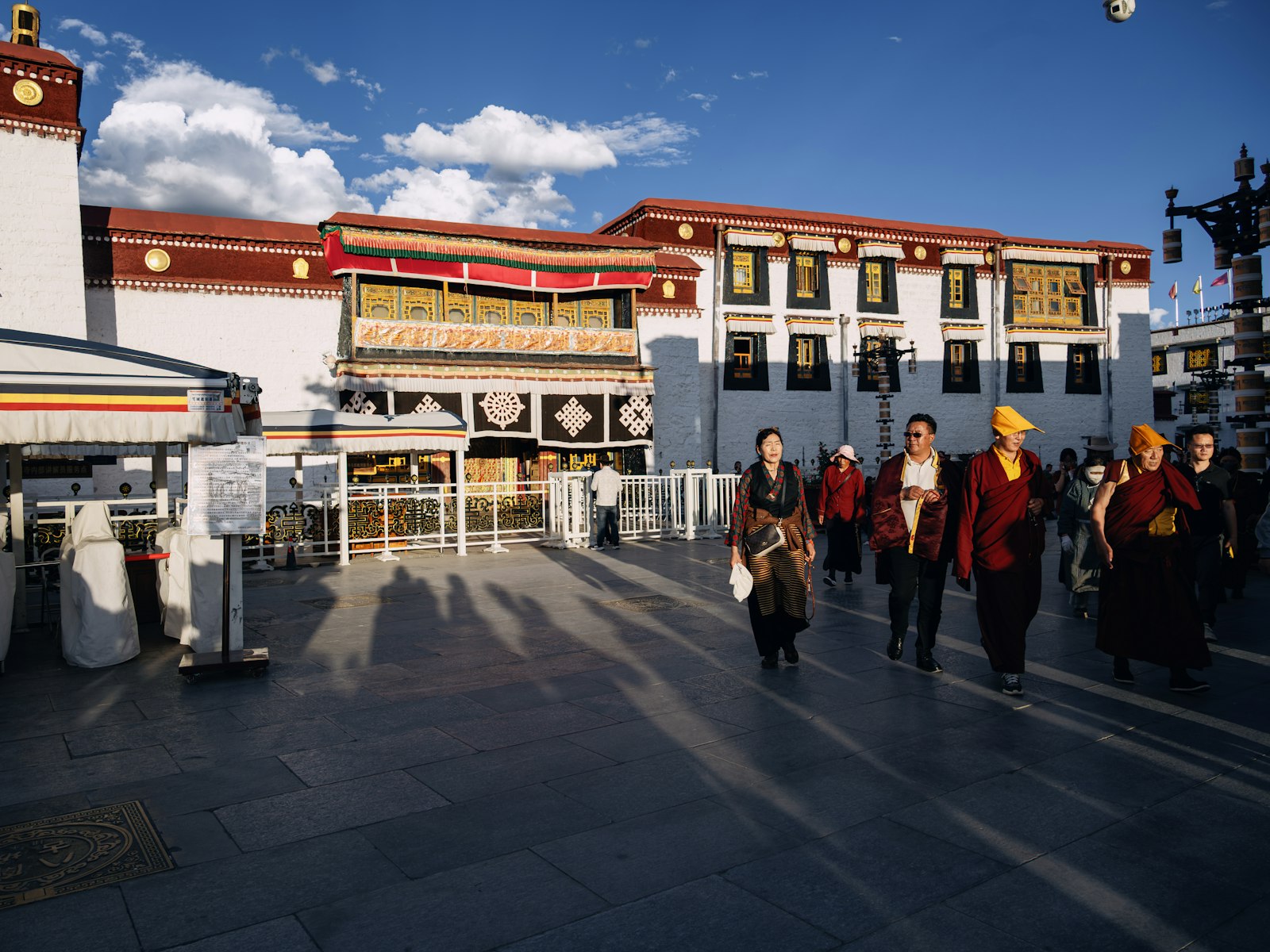 Pilgrims and maroon-robed monks walking across a sunlit flagstone square in front of a whitewashed Tibetan temple building, long shadows stretching over the stones