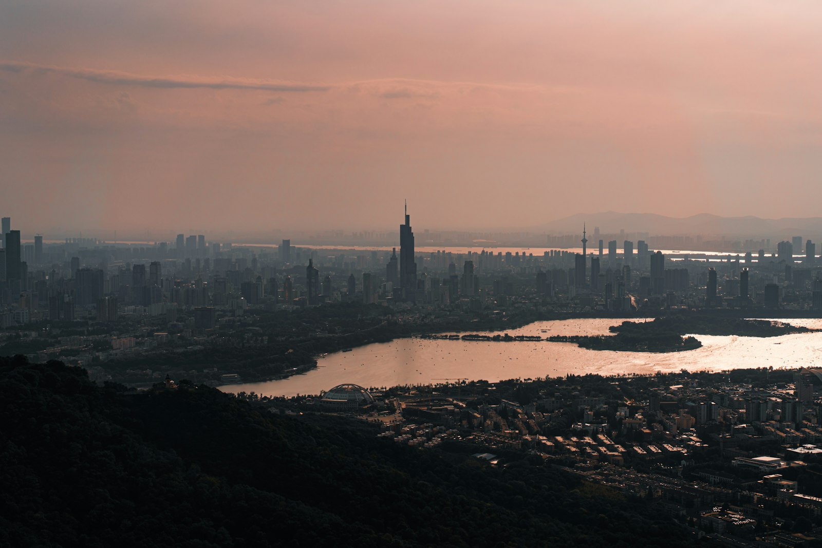 Nanjing skyline at dusk — the Ming City Wall curves past Xuanwu Lake toward Purple Mountain, city lights beginning to glow