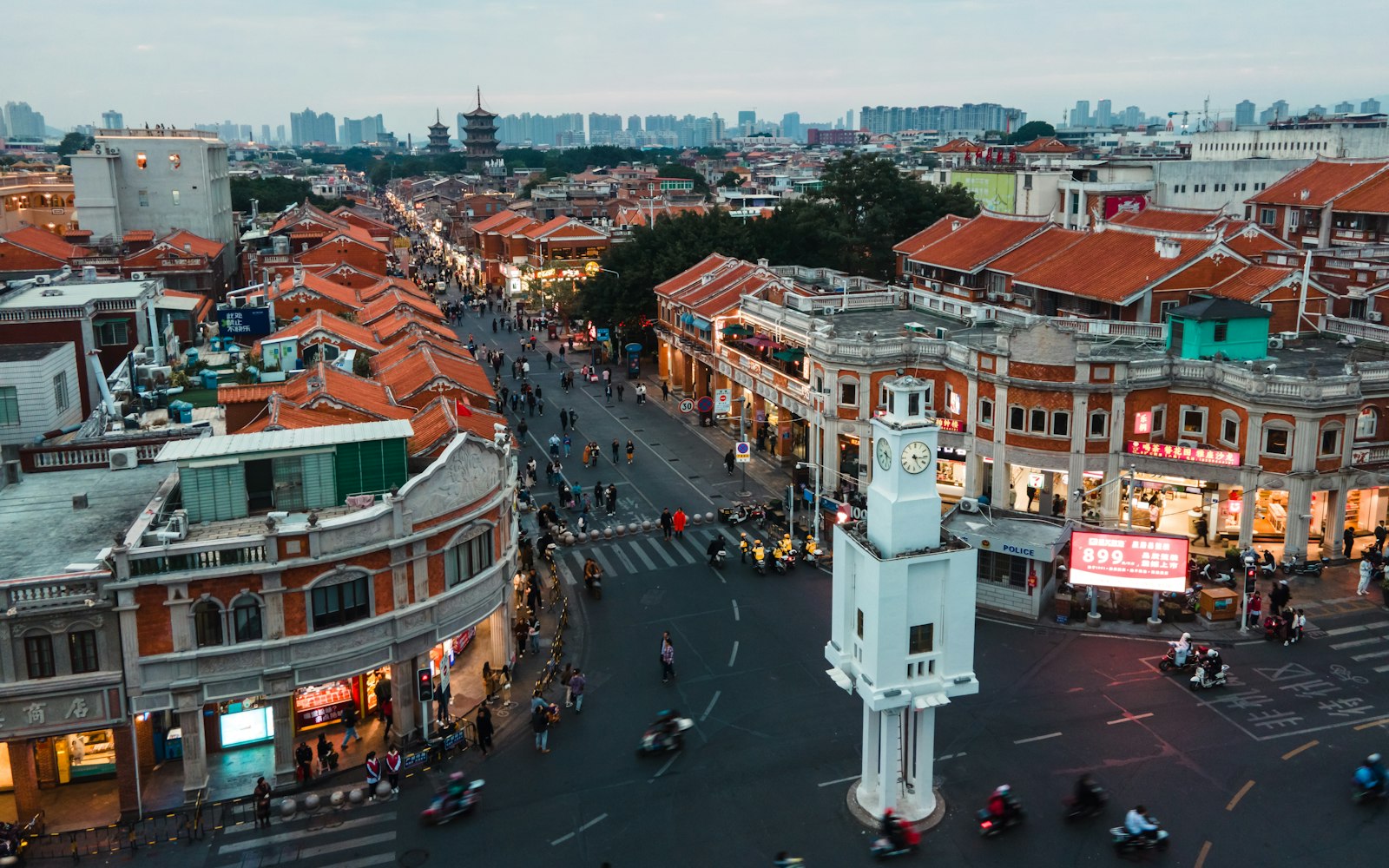 Aerial view at dusk of a white clock tower at a busy old-town junction in Quanzhou, arcaded red-brick shophouses and red-tiled roofs around it, with the twin pagodas of Kaiyuan Temple faint on the horizon