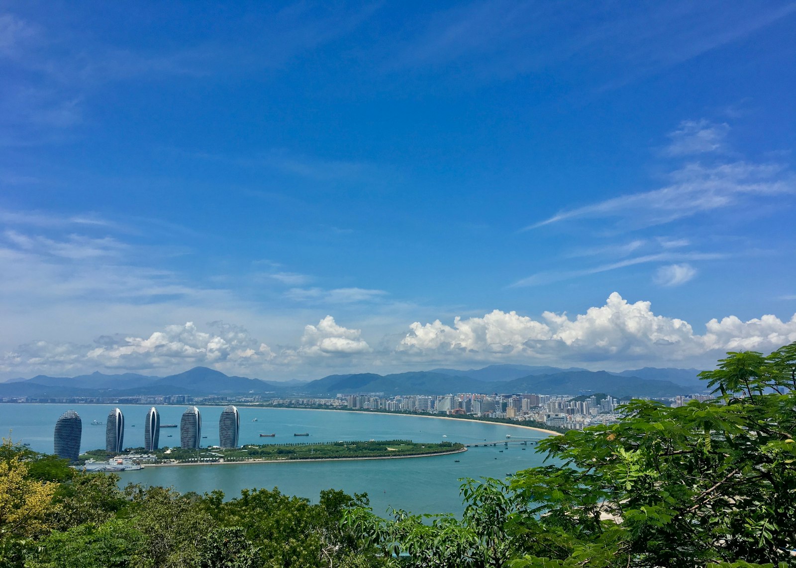 View over Sanya Bay from Luhuitou Park — the curved white towers of Phoenix Island on a green spit, the long crescent of the city beach and mountains behind, under a deep blue sky