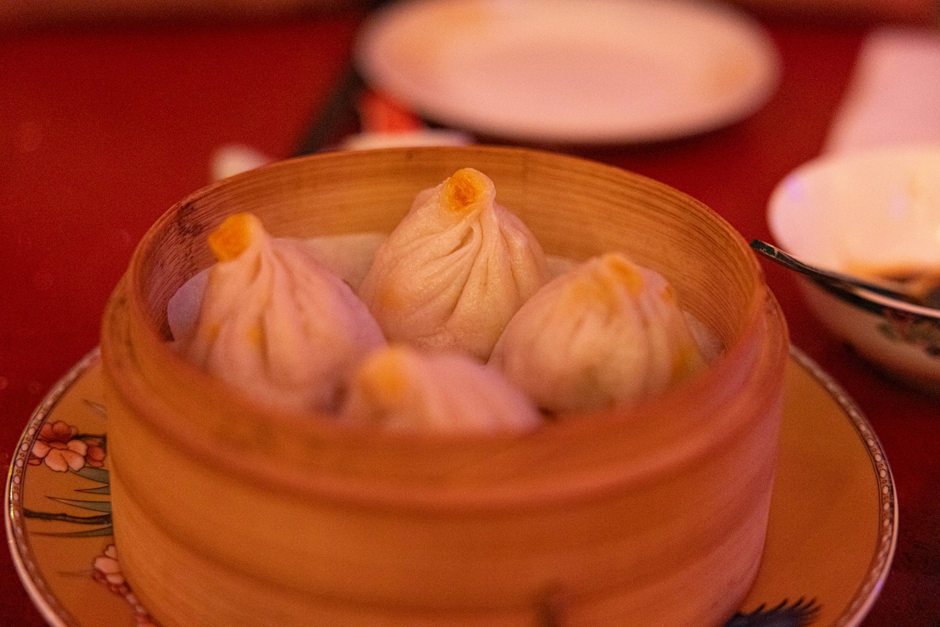 A bamboo steamer of xiaolongbao soup dumplings, one lifted by chopsticks onto a soup spoon, ginger-and-vinegar dish beside it
