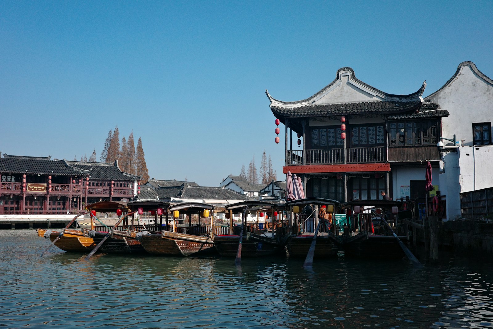 Wooden gondola-style boats moored on the canal at Zhujiajiao beside old teahouse buildings with upturned gables and red lanterns, under a clear blue sky