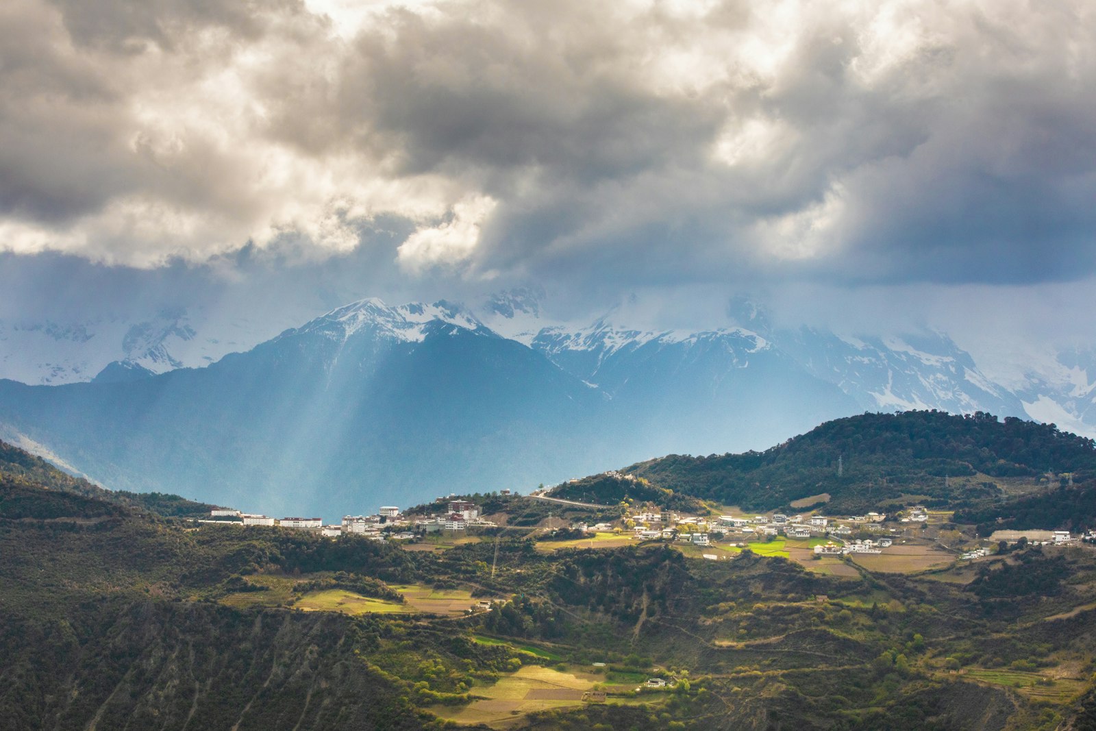 Shafts of sunlight breaking through heavy cloud onto a hillside settlement, the snow peaks of the Meili range rising in the haze behind