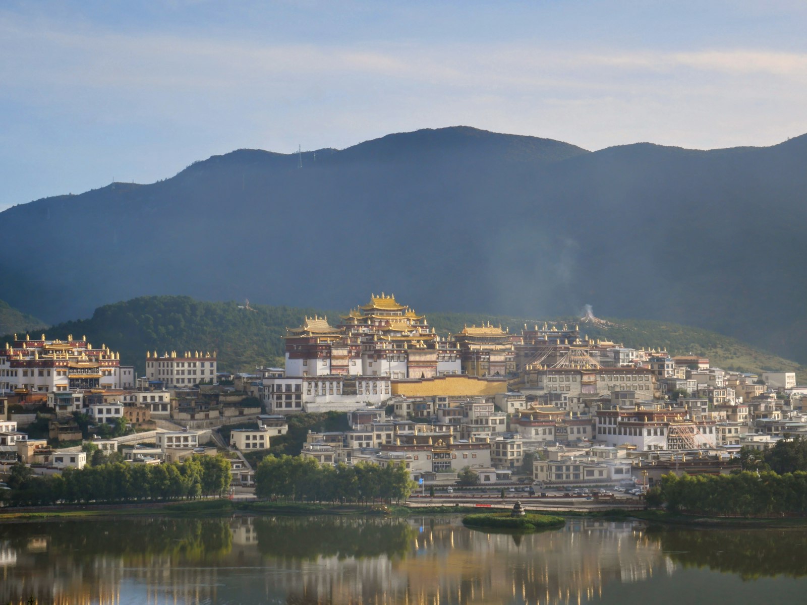 Ganden Sumtseling (Songzanlin) Monastery in soft golden-hour light — gilded temple roofs and white halls on the hillside above a lake, houses at its foot and dark ridgelines behind