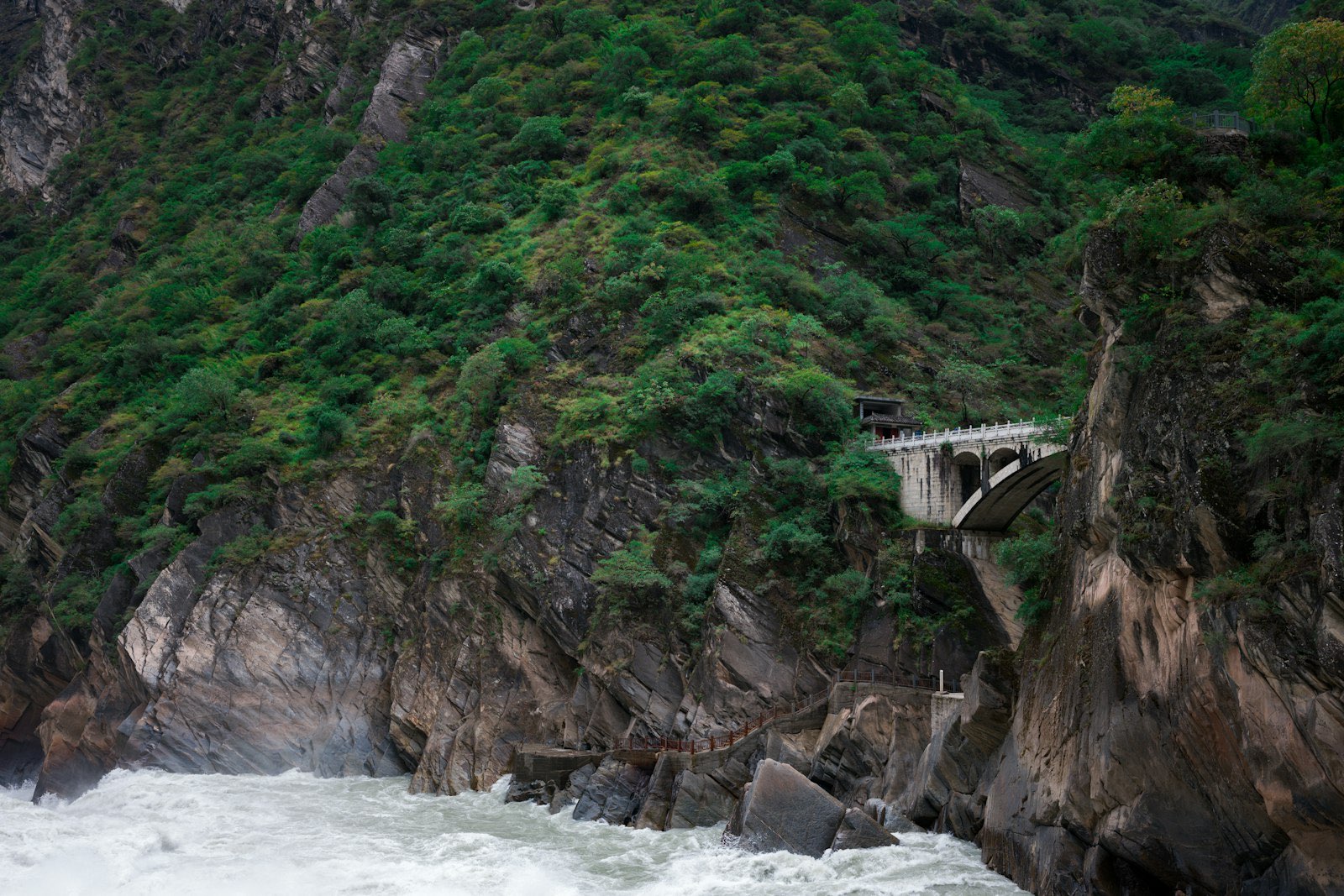 Tiger Leaping Gorge: the Jinsha River churning white below steep, forested cliffs, a stone viaduct and a railed path clinging to the rock face