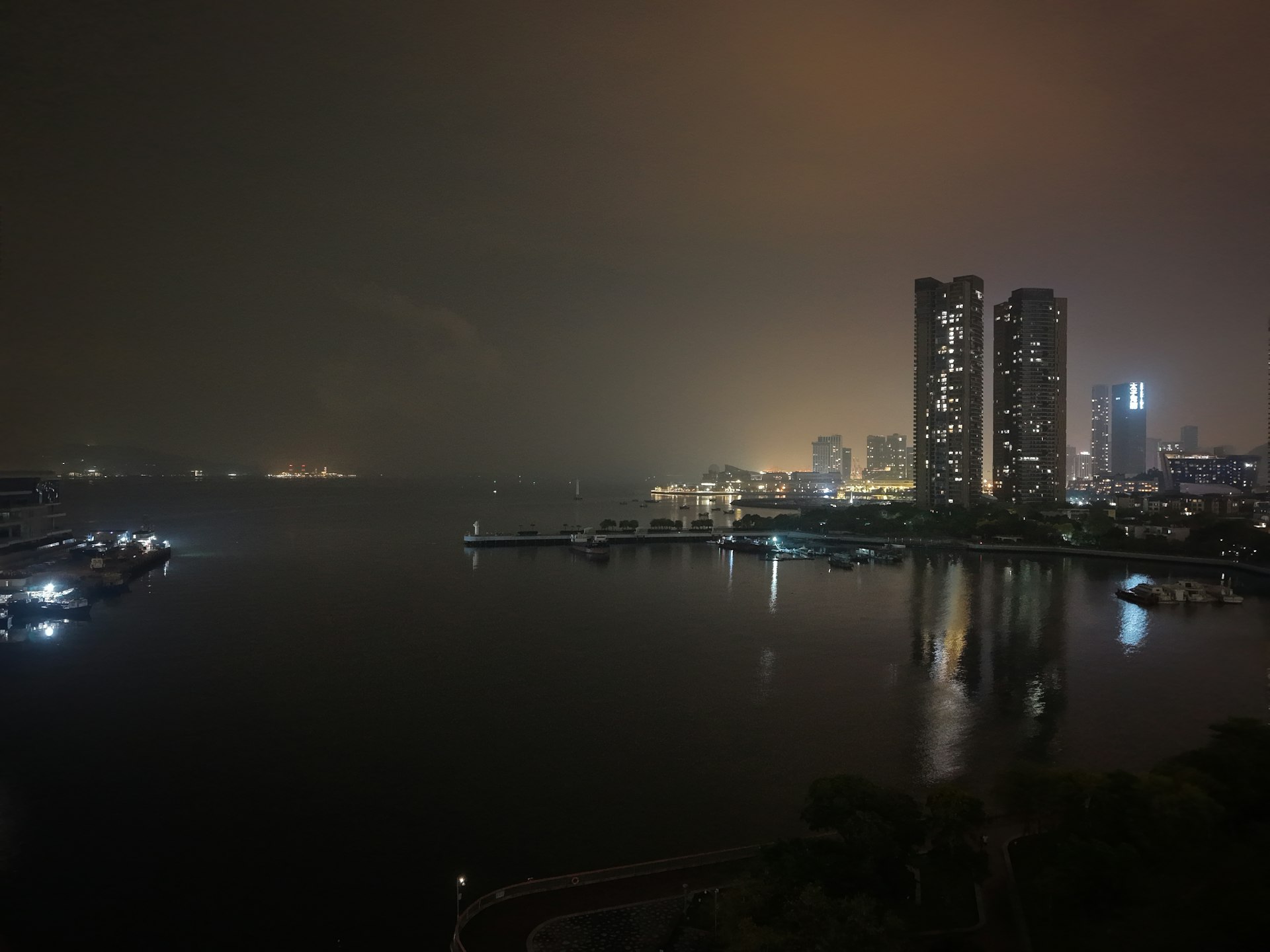 Shenzhen Bay Park boardwalk at golden hour — joggers, palm trees, and the Hong Kong skyline hazy across the water