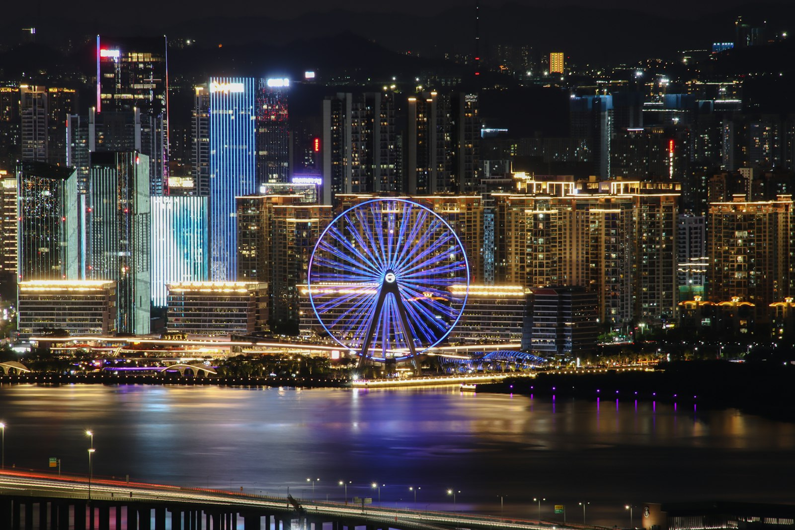 A brightly lit Ferris wheel among glowing waterfront towers, mirrored in the bay at night, with light trails on a bridge in the foreground