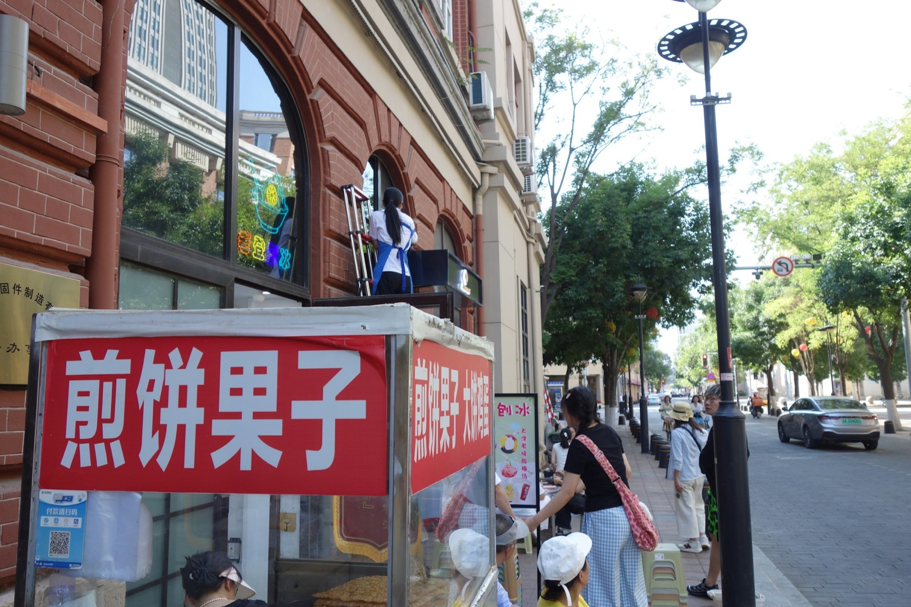 A street stall with a red sign reading jianbing guozi in Chinese characters, beside a red-brick building with arched windows