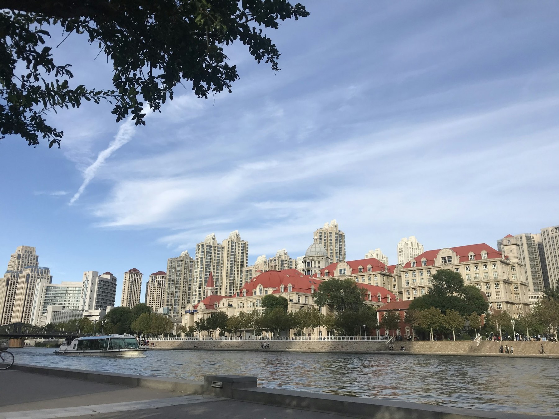 Red-roofed European-style buildings and apartment towers along the Hai River under a blue sky, with a boat on the water