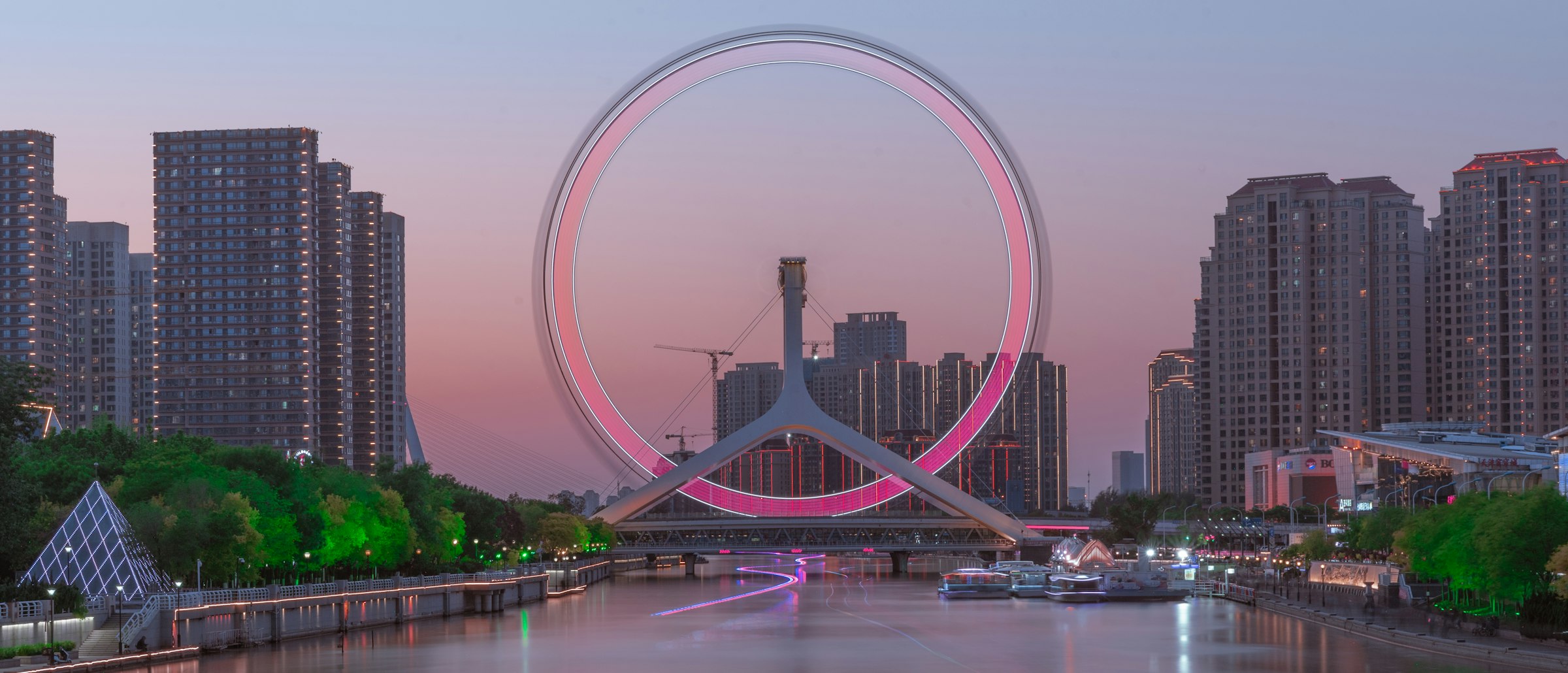The Tianjin Eye Ferris wheel lit pink at dusk on a bridge over a calm river, with apartment towers on both banks