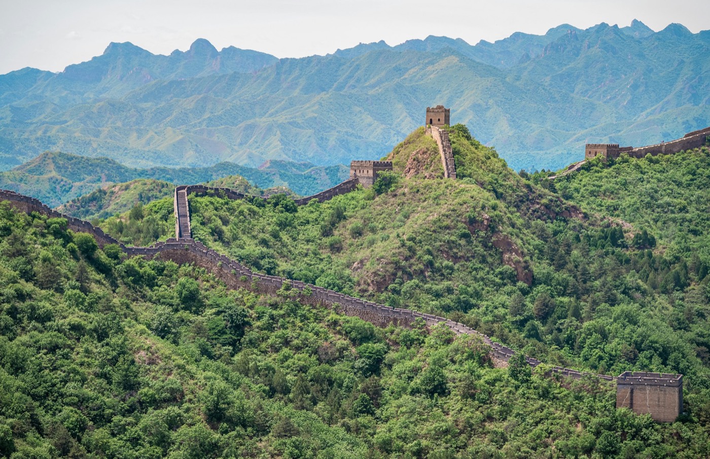 A stone section of the Great Wall with watchtowers climbing a green ridge, with hazy blue mountains behind