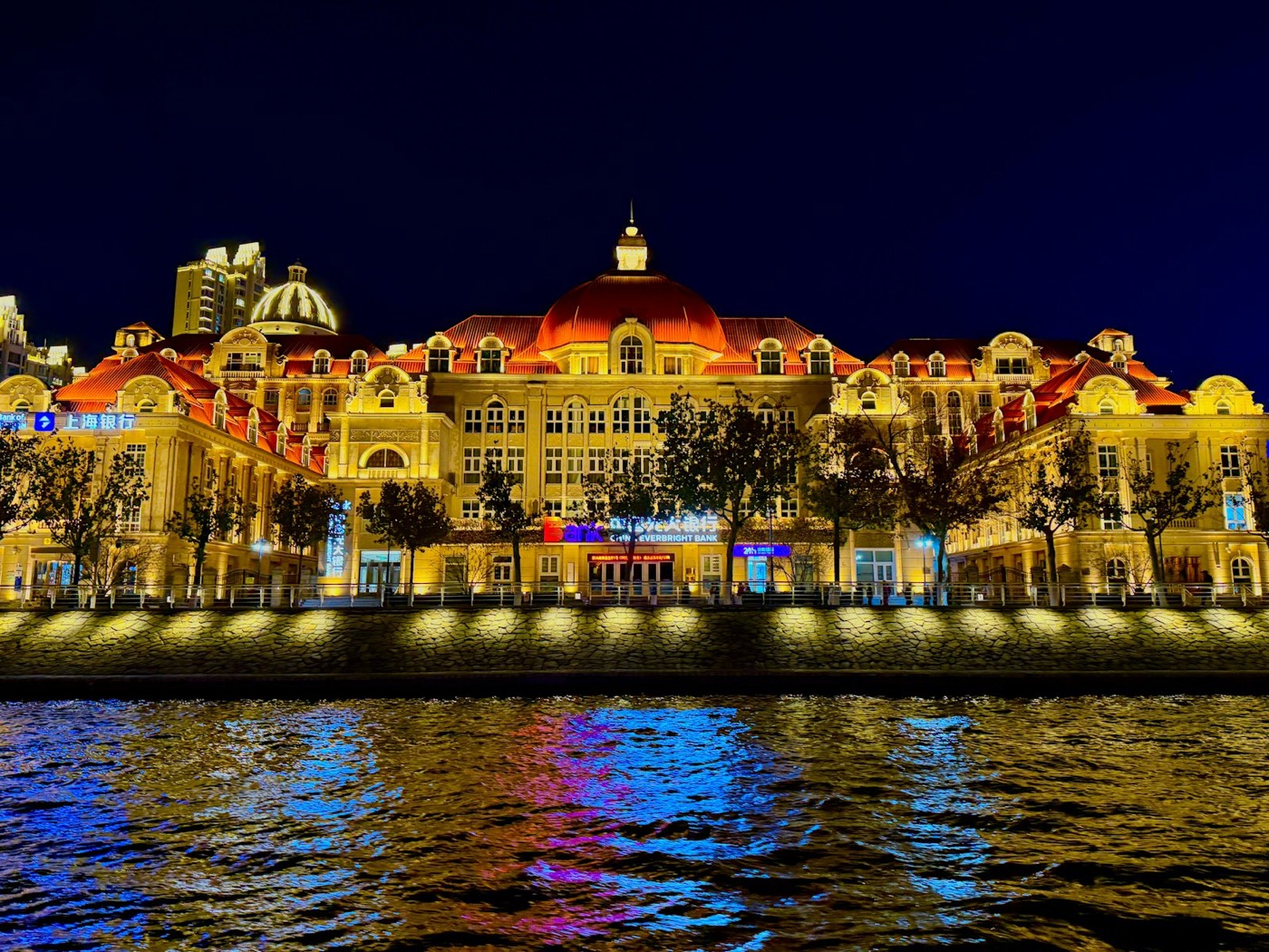 A European-style building with a red domed roof lit in gold at night, with colorful lights reflected in the river