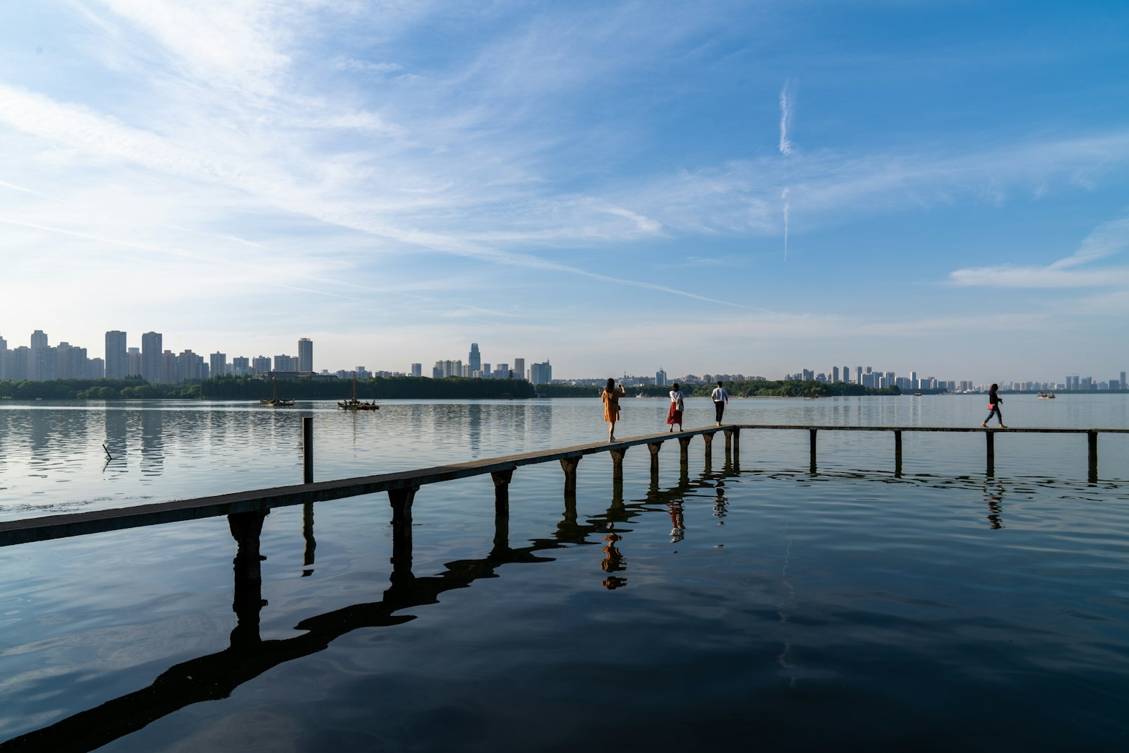 Walkers on a narrow boardwalk stretching out over the calm, blue water of East Lake, Wuhan, with the city skyline across the lake