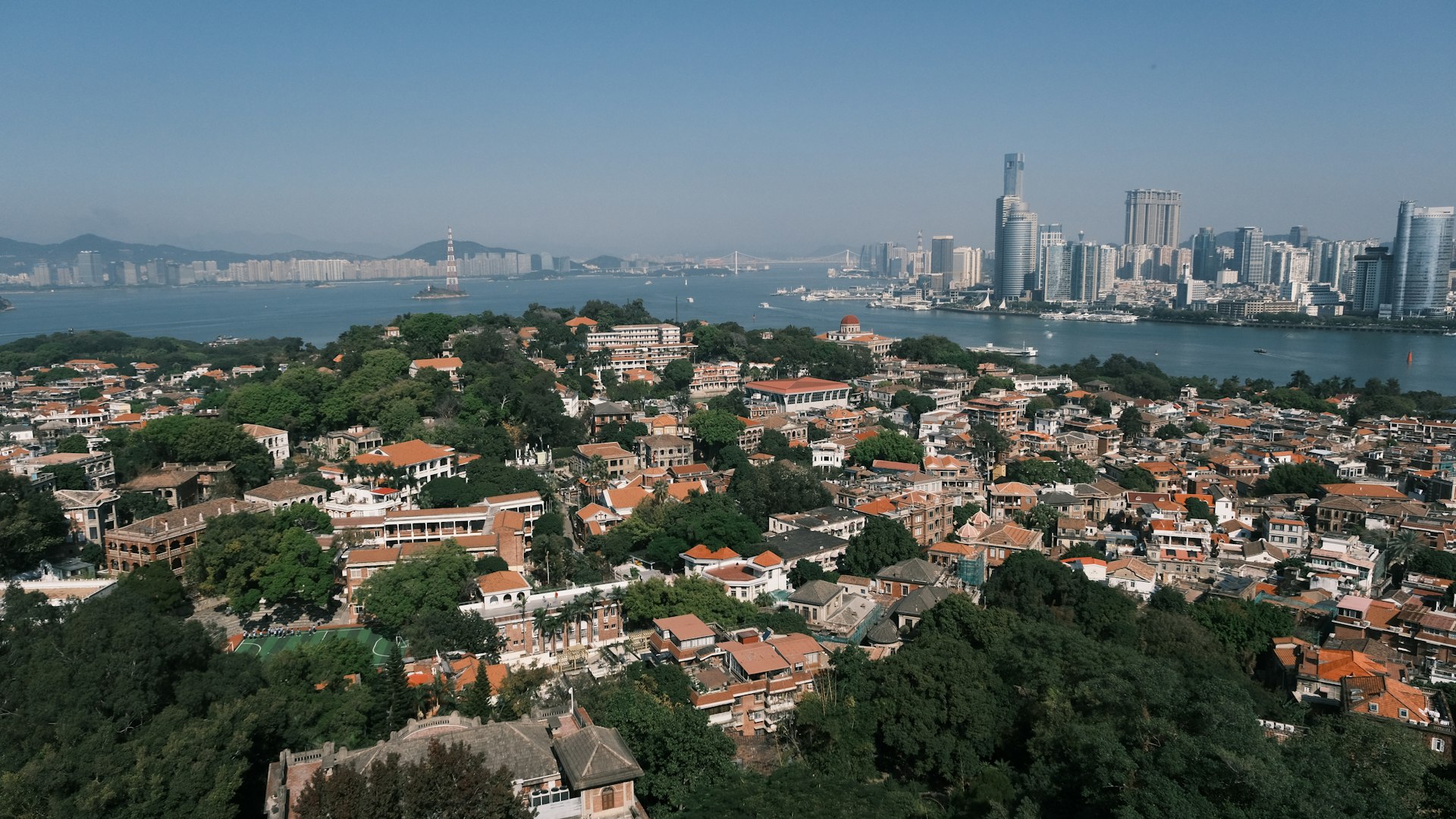 Gulangyu Island's colonial villas and red rooftops above the sea, with Xiamen's skyline across the water — hero shot