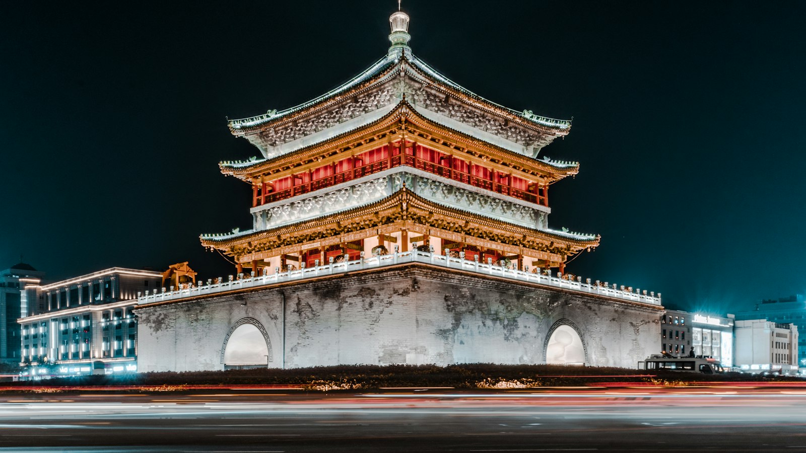 Xi'an's Bell Tower lit up at night — three tiers of golden eaves and red galleries on a grey brick base, with streaks of car headlights in front