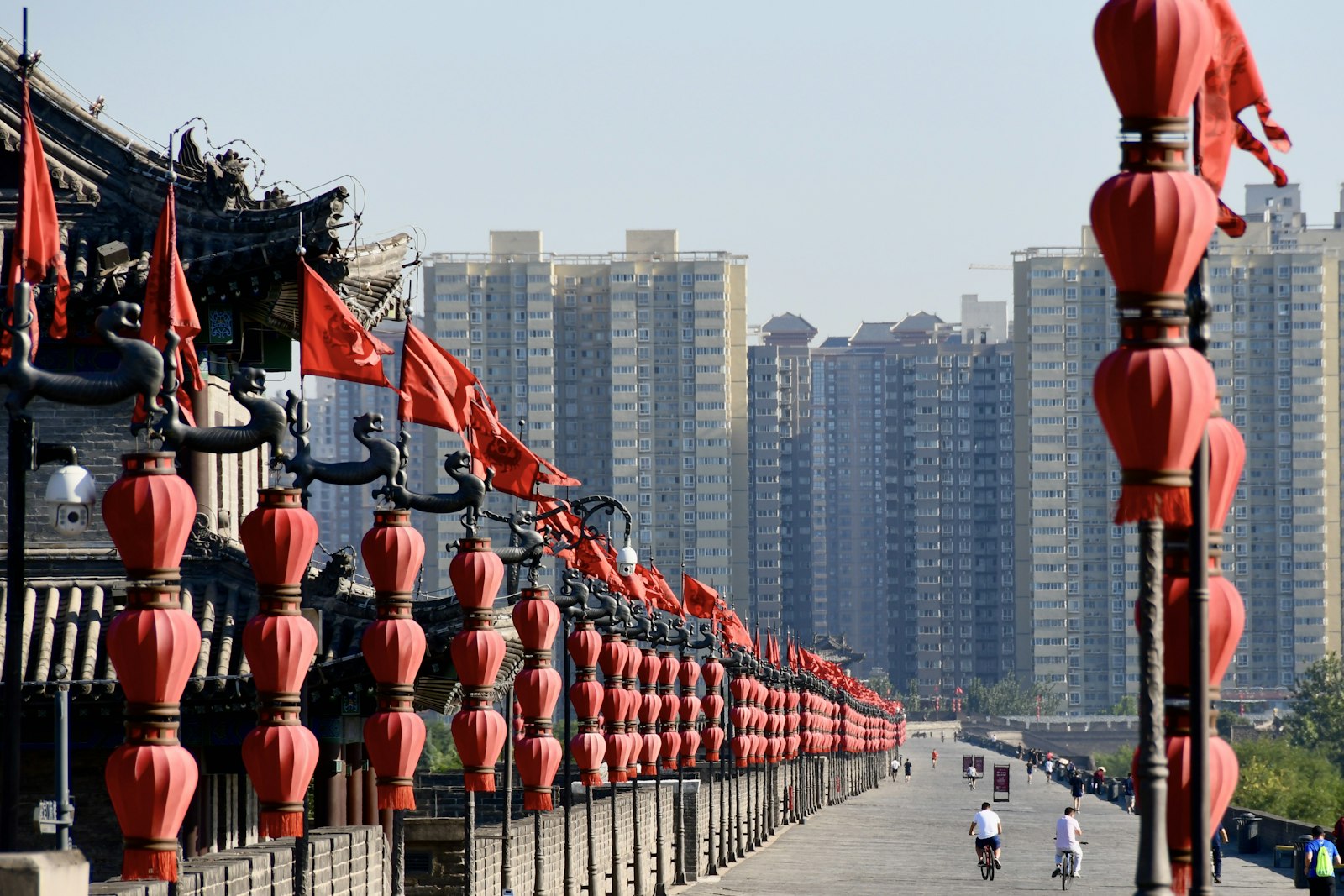 Rows of red lanterns and flags lining the top of Xi'an's city wall, with two cyclists riding the wide brick walkway toward apartment towers in the distance
