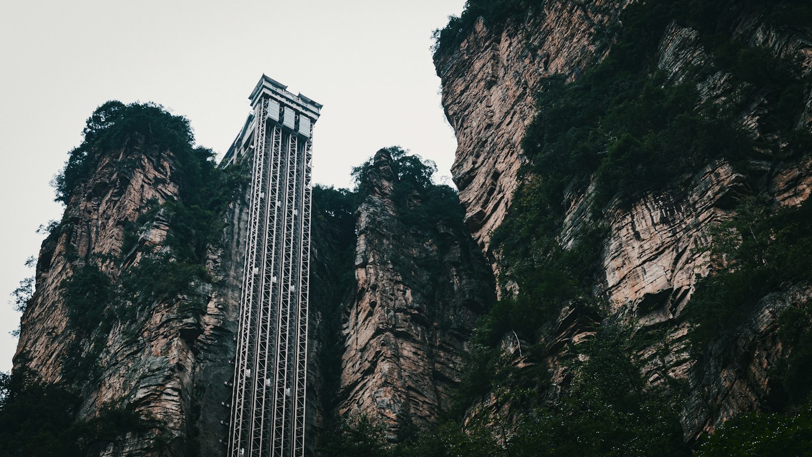 The Bailong Elevator seen from below — a steel-framed lift tower climbing a sheer sandstone cliff between forested rock walls
