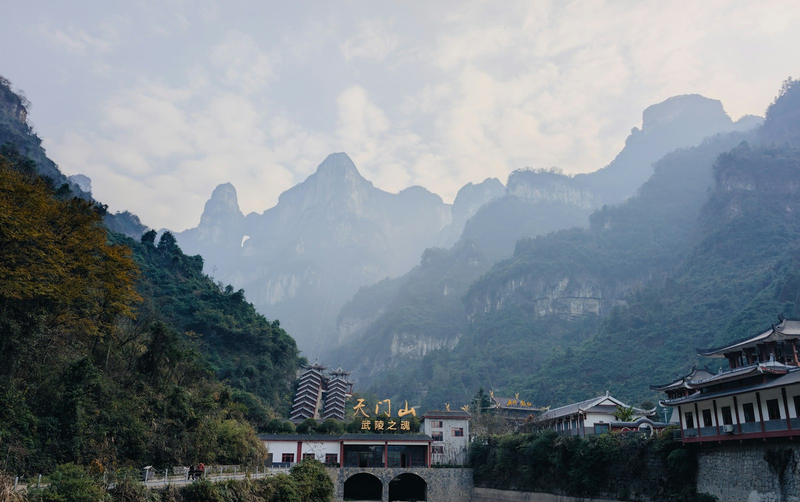 Tianmen Mountain from its foot — a gold 天门山 signboard and pagoda-roofed buildings below misty cliff-walled peaks, with the sky showing through the arch of Heaven's Gate in the ridge